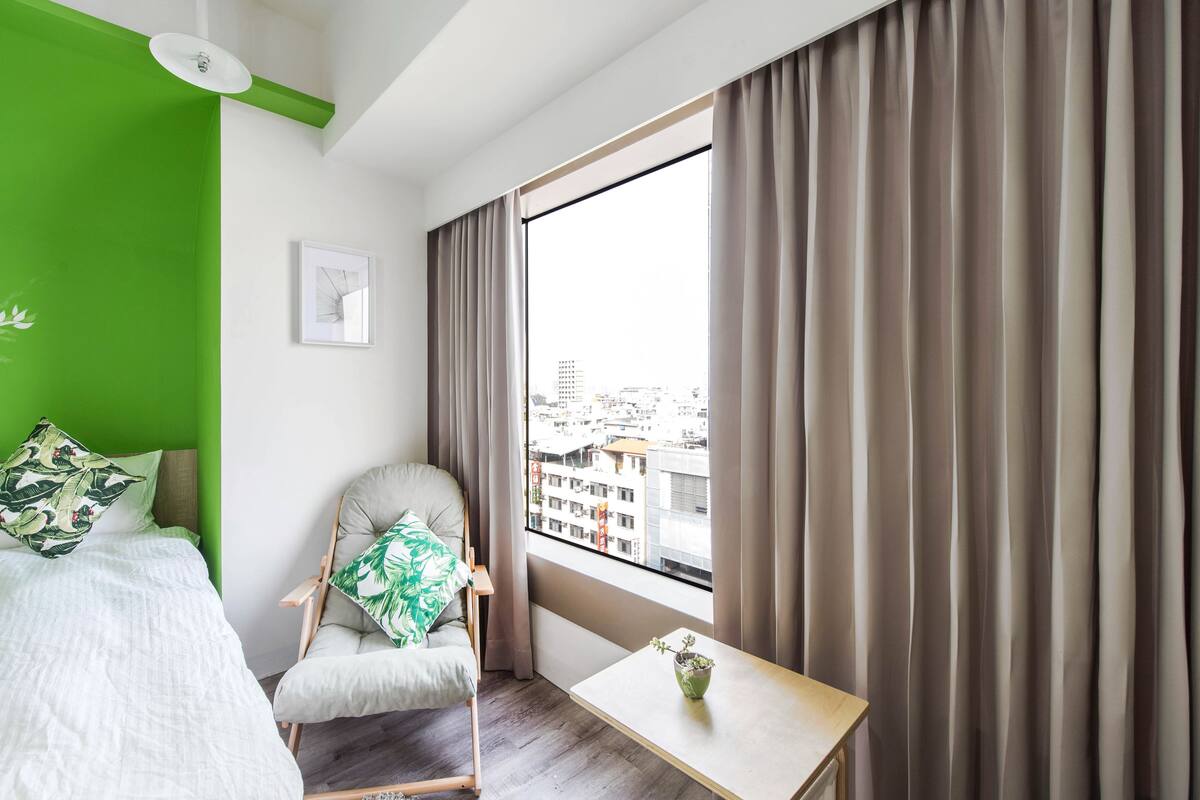 A modern hotel room with a green accent wall, a large window with curtains, a bed with green and white bedding, a chair with a green pillow, and a small table with a plant.