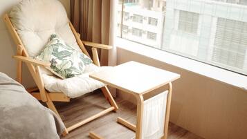 A modern room with a wooden floor, a cushioned chair with a floral pillow, a small wooden table, and a view of buildings through a window.
