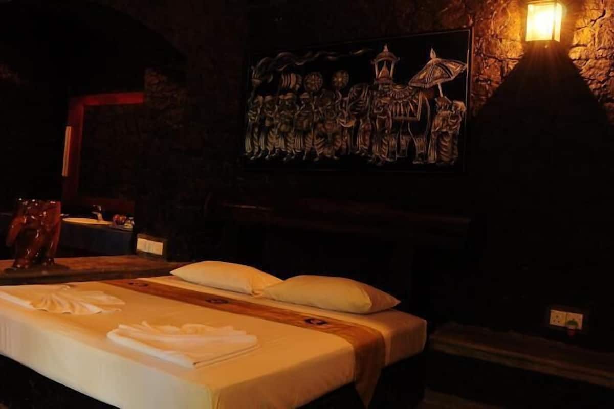 A dimly lit room with a bed, a wooden headboard, a wall-mounted light, and a framed artwork above the bed.
