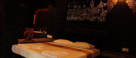 A dimly lit room with a bed, a wooden headboard, a wall-mounted light, and a framed artwork above the bed.