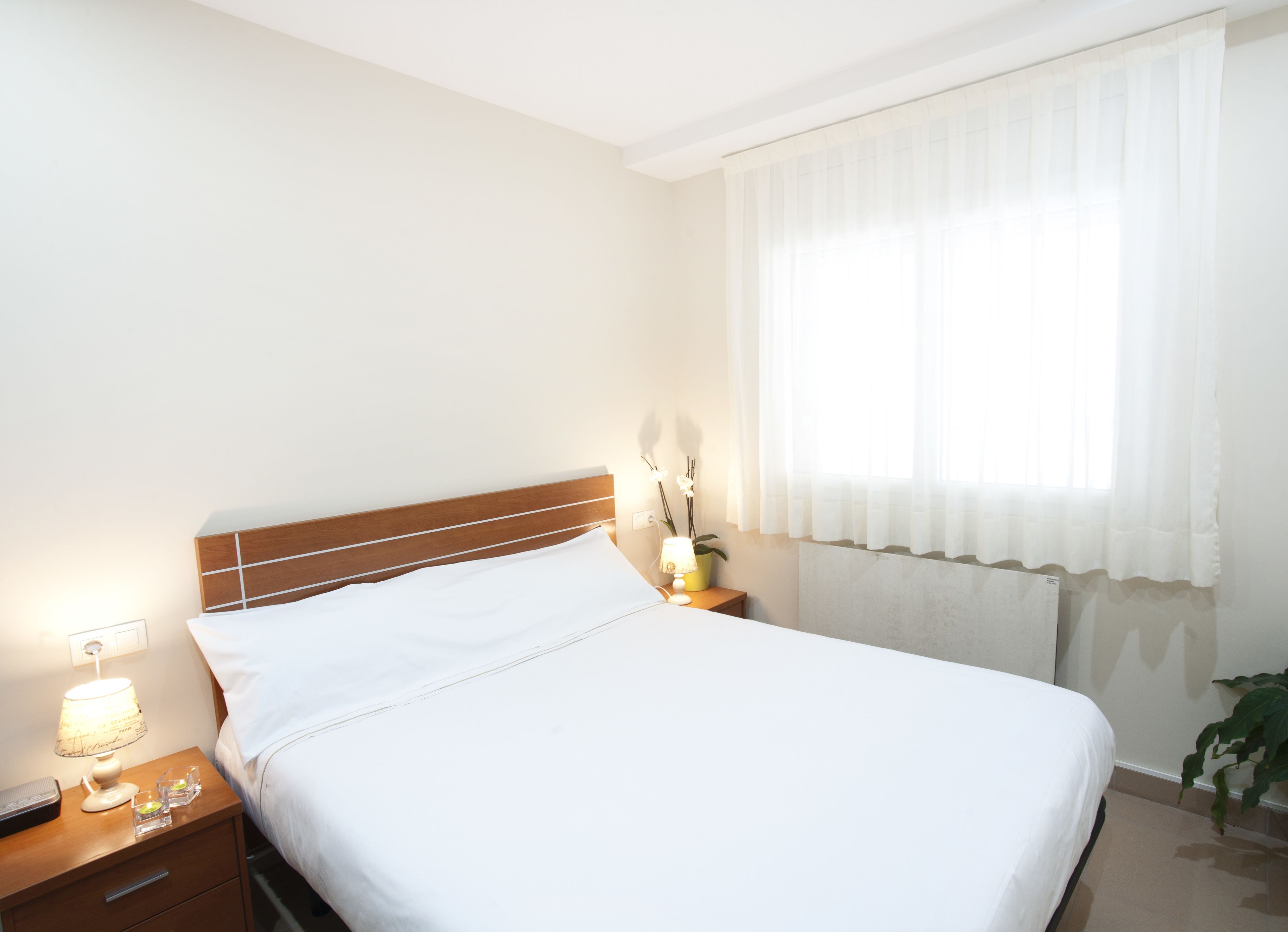 A neatly made bed with white linens, a wooden headboard, a bedside lamp, a small table with a lamp and a plant, and a window with sheer curtains.