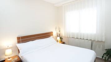 A neatly made bed with white linens, a wooden headboard, a bedside lamp, a small table with a lamp and a plant, and a window with sheer curtains.