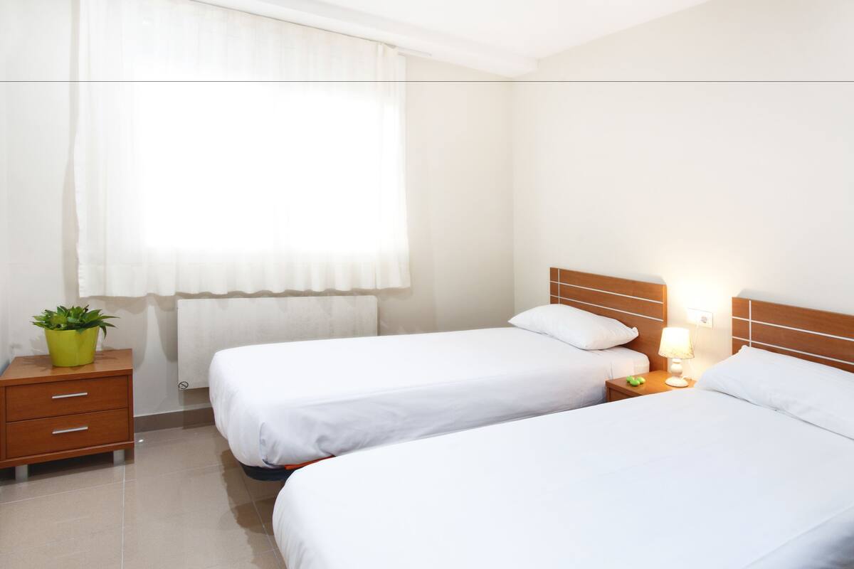 Habitación de hotel con dos camas individuales, una mesita de noche con lámpara, una cómoda pequeña y una ventana con cortinas.