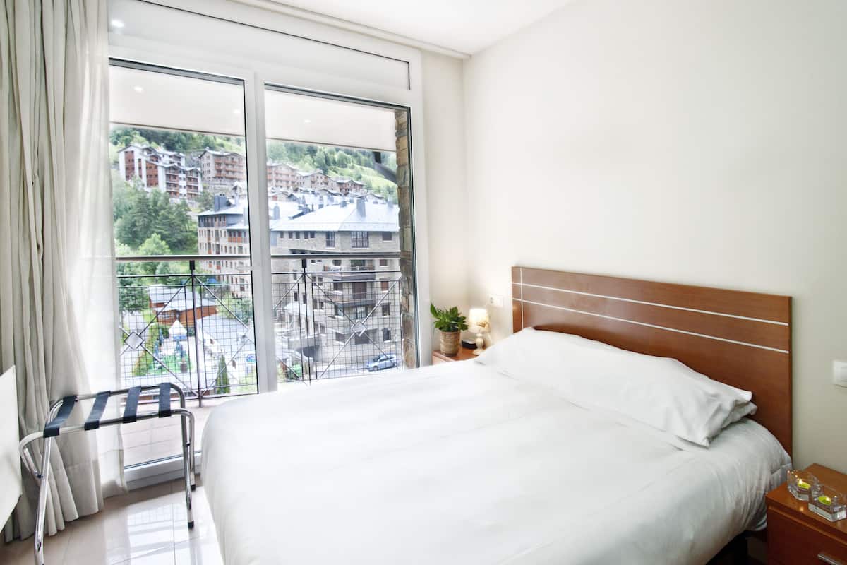 Habitación de hotel con una cama grande, vistas a edificios a través de una ventana, una mesita de noche con lámpara y una silla de ruedas.