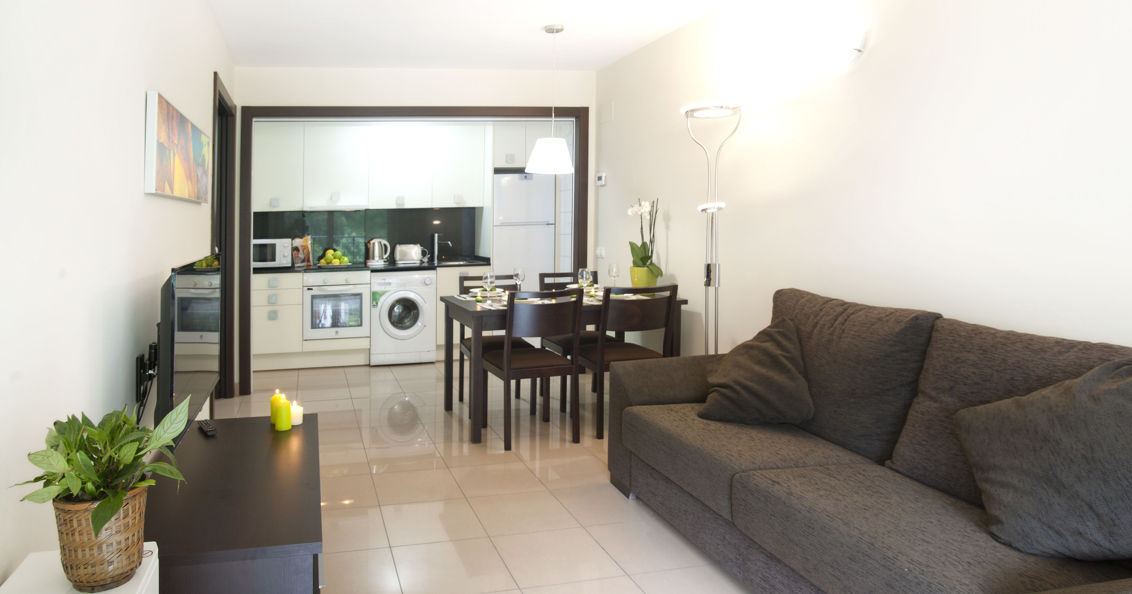 Una sala moderna con un sofá gris, una mesa de comedor con sillas, una mesita auxiliar con una planta y una cocina al fondo.