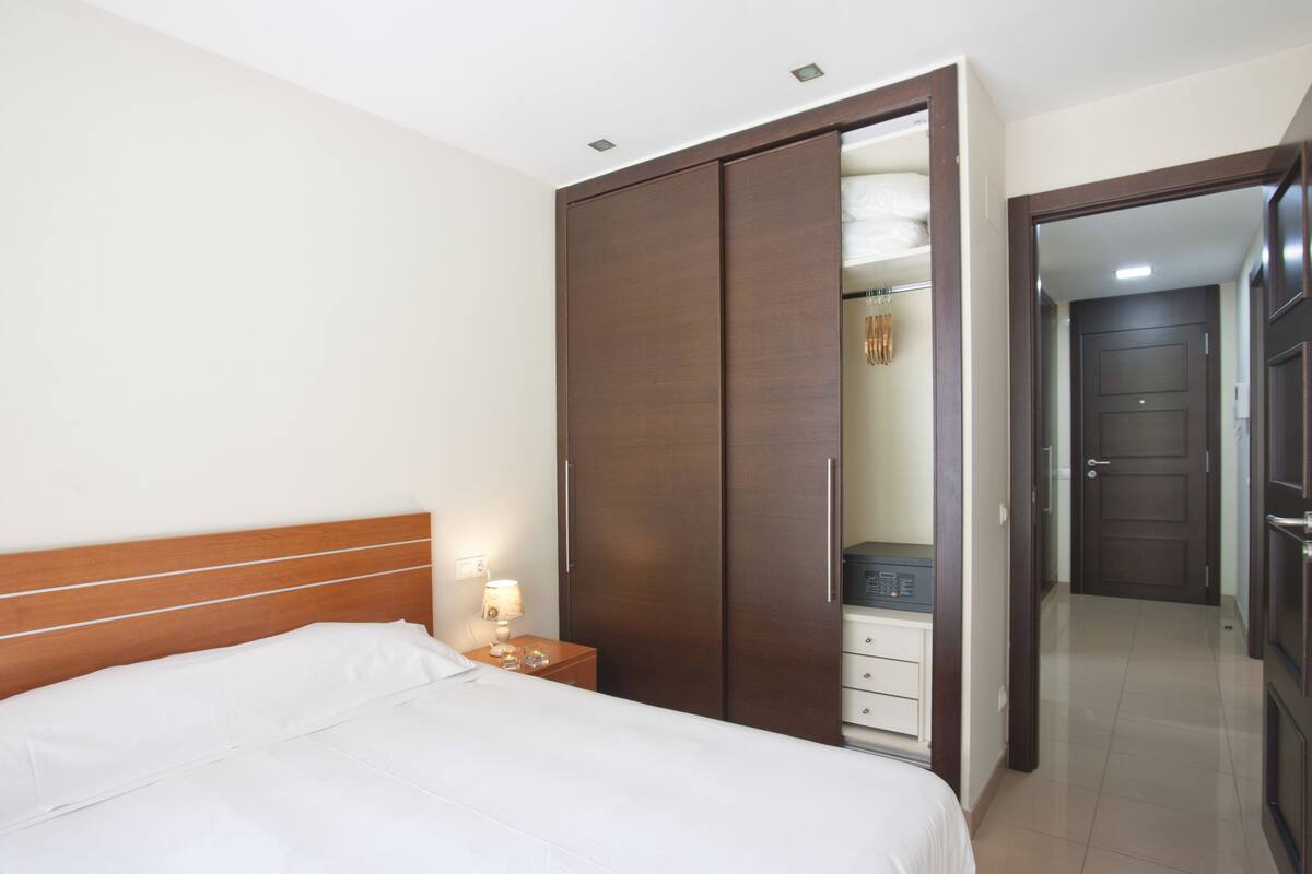 Un dormitorio con cama, un armario de madera, una mesita de noche con lámpara, y una puerta que da a otra habitación.