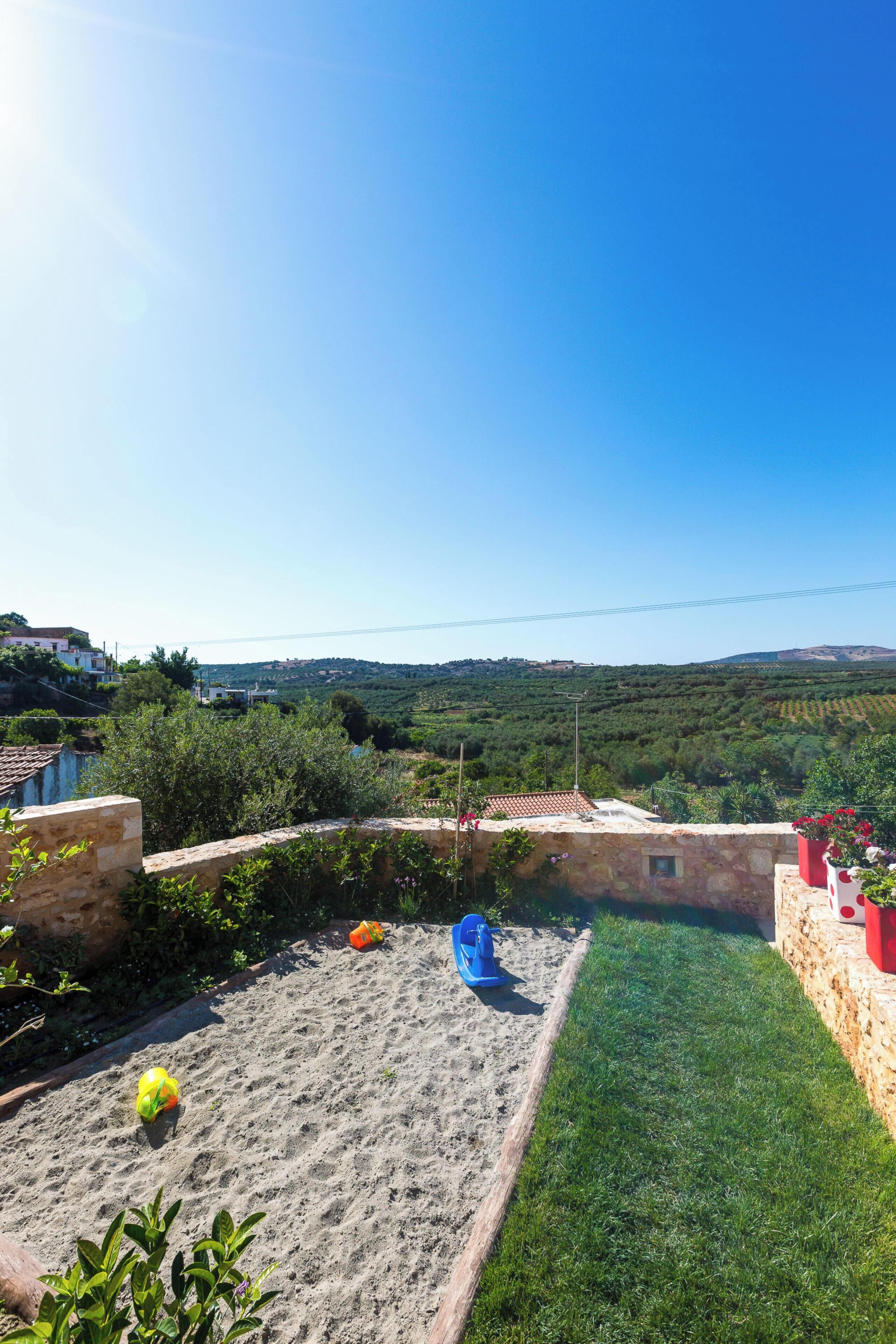 A backyard with a sandy play area, a blue slide, and a view of distant hills.