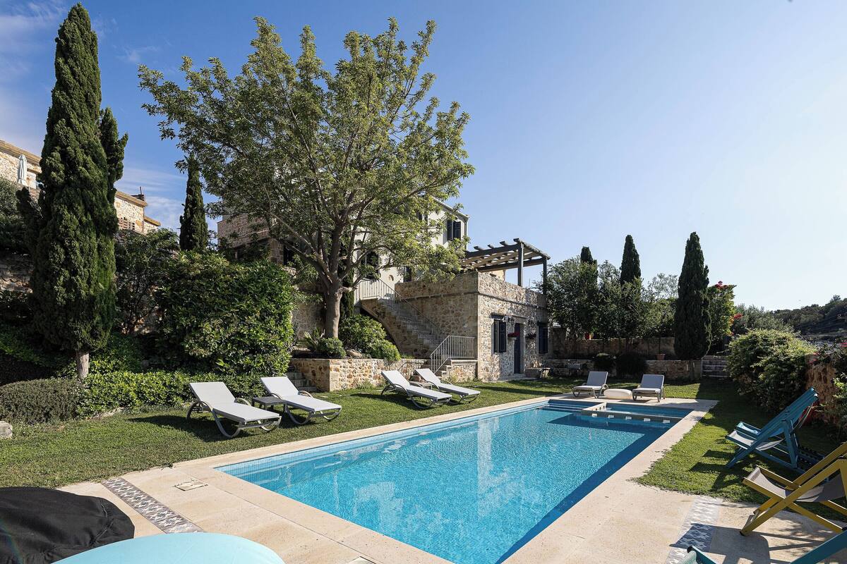 Une piscine entourée de chaises longues, d’une pergola et de bâtiments en pierre en arrière-plan.