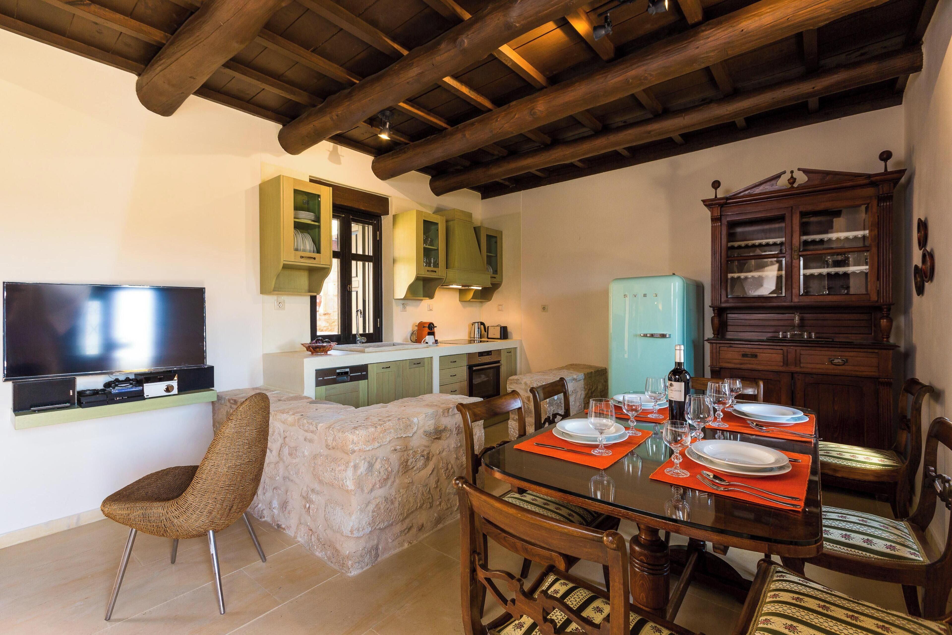 Un comedor con una mesa dispuesta para cuatro personas, un televisor montado en la pared y una zona de cocina con armarios y un refrigerador.