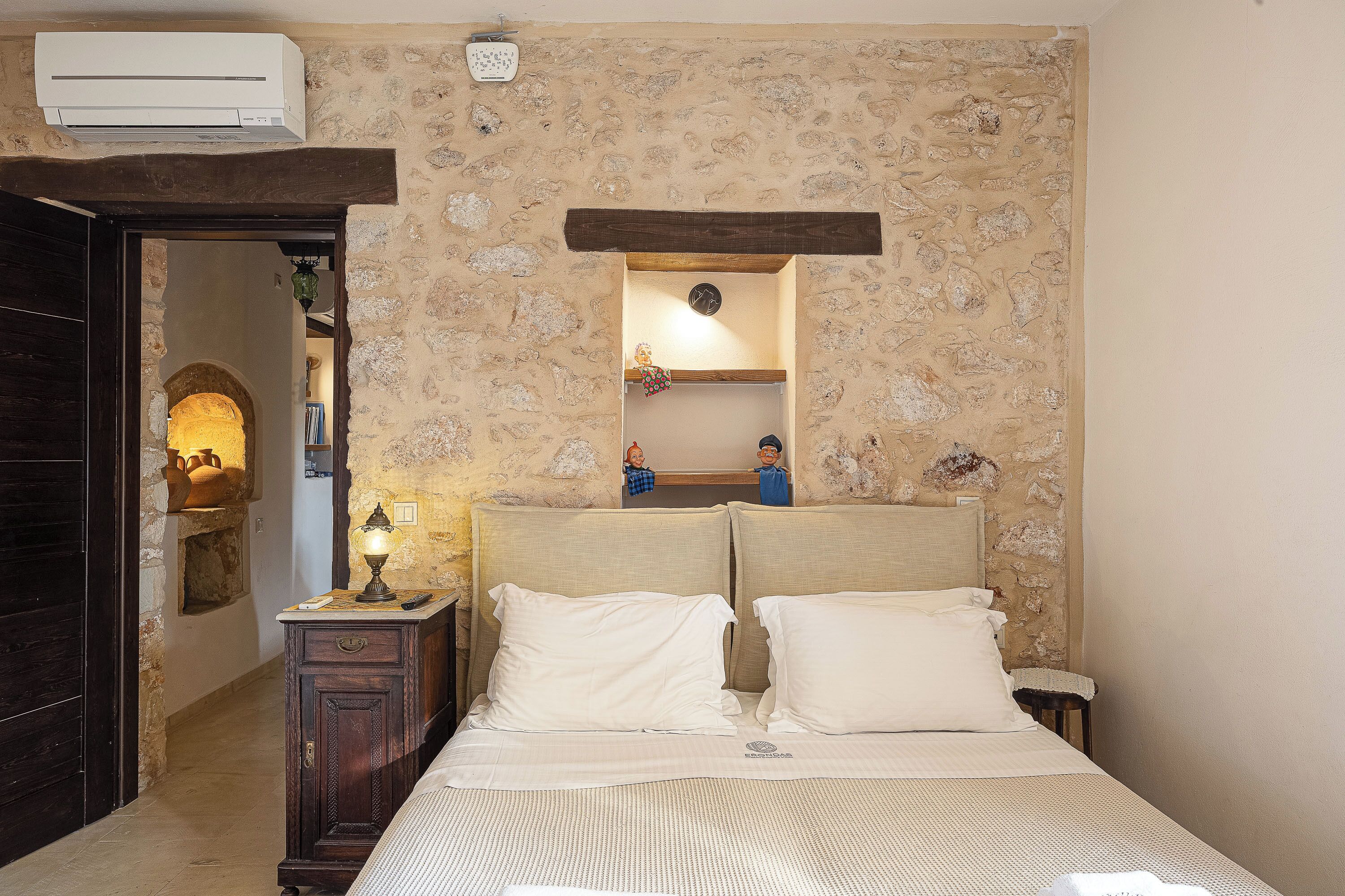 A bedroom with a stone wall, a bed with white bedding, a nightstand with a lamp, and a wooden door.