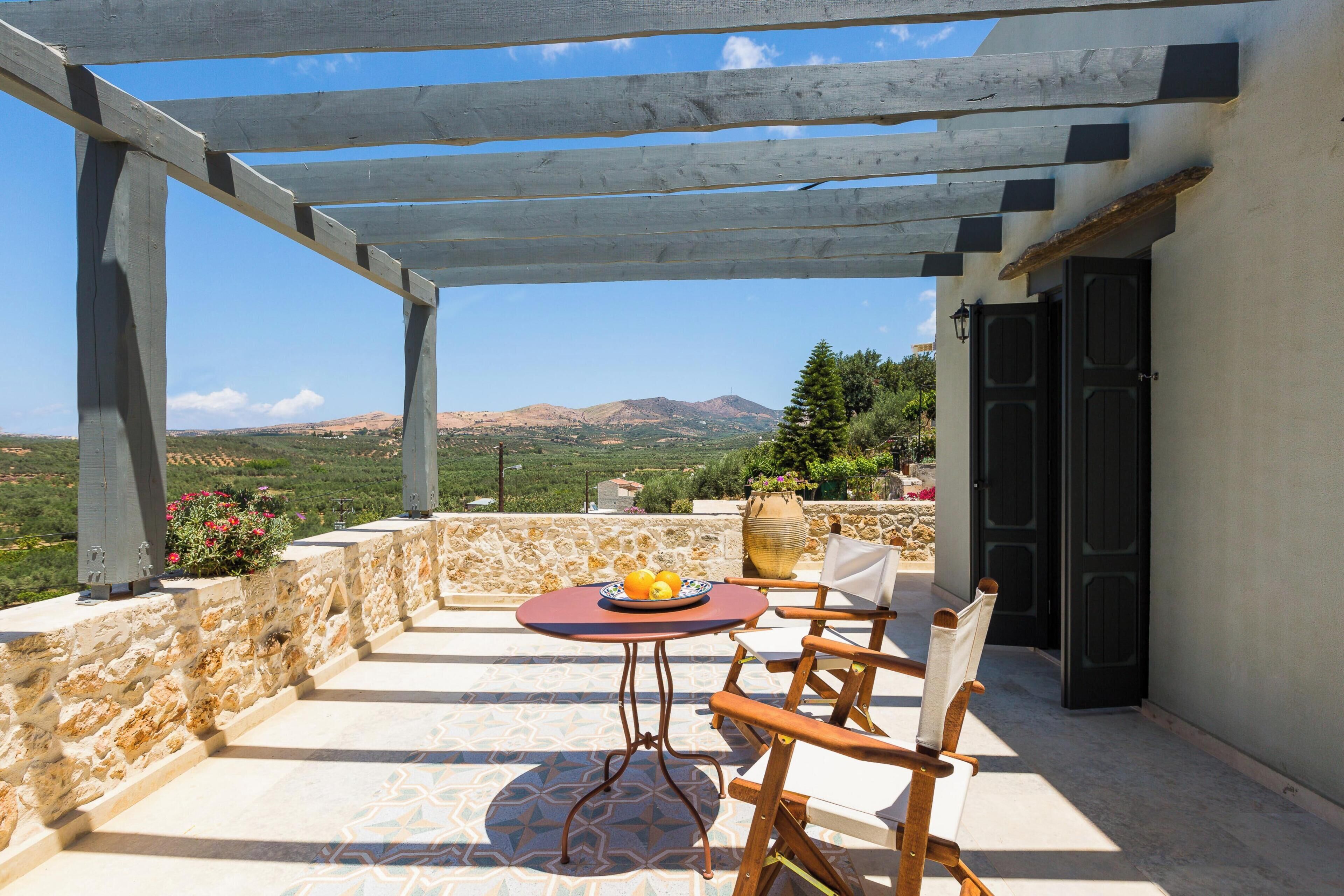 A covered outdoor seating area with a table and chairs, a stone wall, and a view of the landscape.