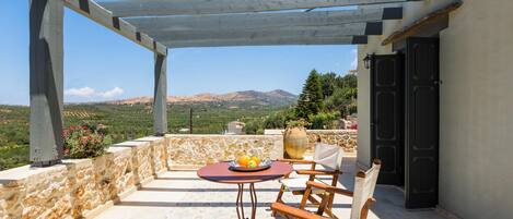 A covered outdoor seating area with a table and chairs, a stone wall, and a view of the landscape.