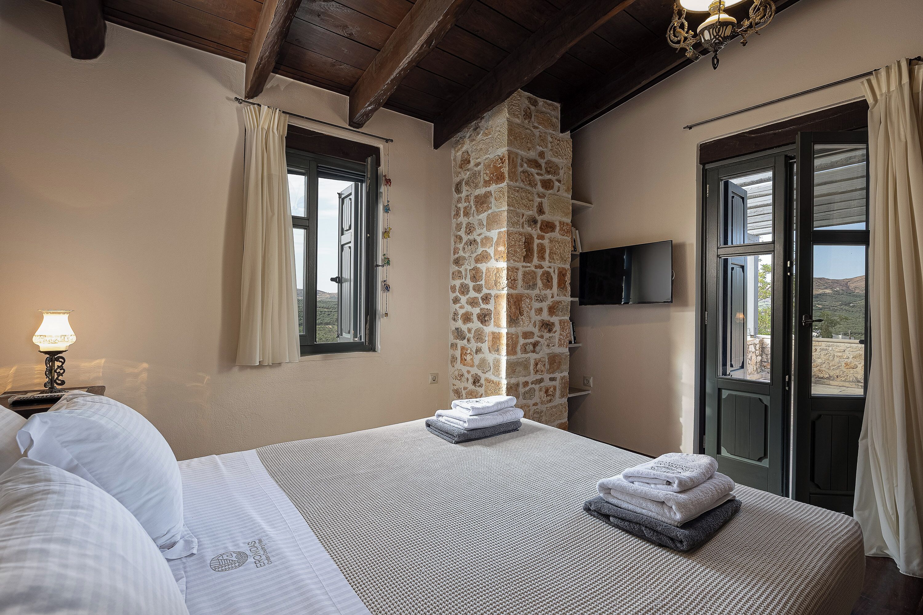Un dormitorio con un pilar de piedra, una cama con sábanas blancas, un televisor en la pared y vistas a través de una puerta abierta.