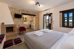 A bedroom with a stone wall, a fireplace, a bed with towels, a chair, and a view through a door. - Erondas Cretan Country Villas (Mylopotamos)