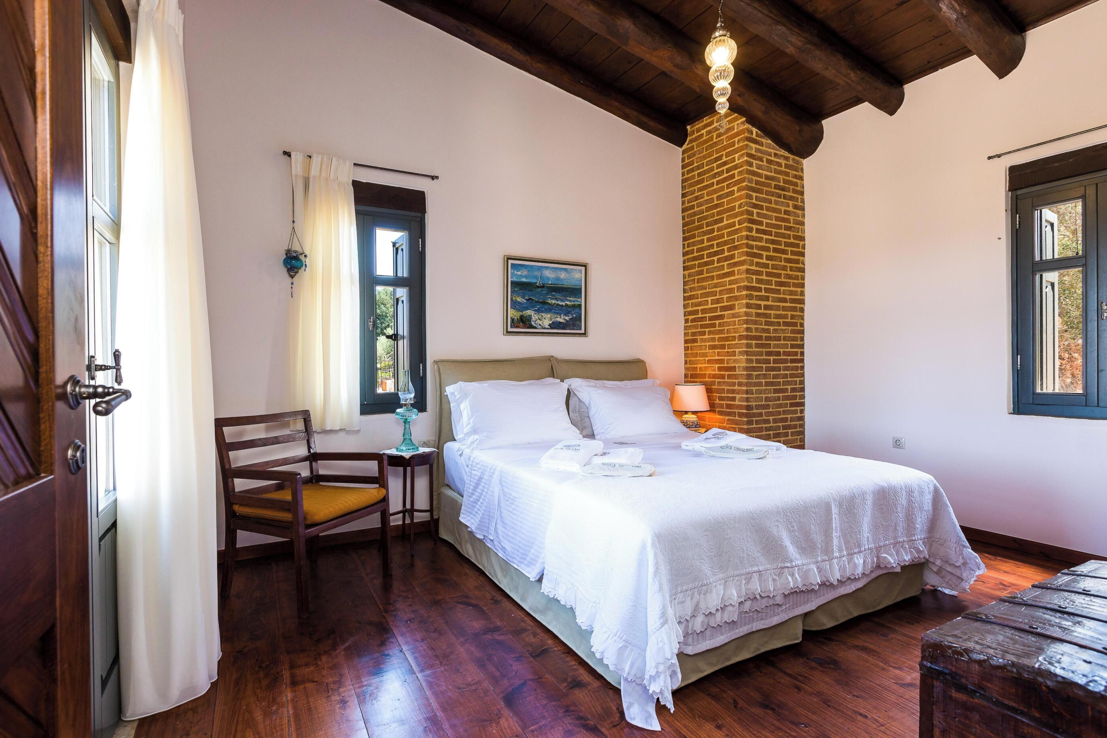 A bedroom with a wooden floor, a bed with white bedding, a chair, a lamp, and a painting on the wall.