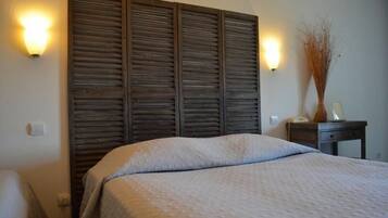 Un dormitorio con una cama grande, cabecera de madera, dos luces fijadas a la pared, una mesita de noche con lámpara y un florero con plantas secas.