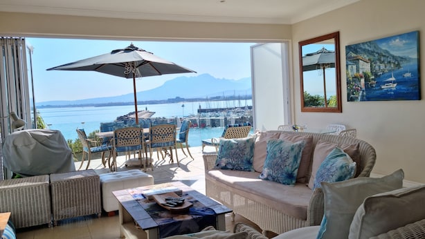 A living room with wicker furniture, a coffee table, and a sofa with cushions. There is a large window offering a view of the sea and boats. - The View (Cape Town)