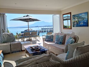 Ein Wohnzimmer mit Korbmöbeln, einem Couchtisch und einer Couch mit Kissen. Ein großes Fenster bietet Blick auf das Meer und Boote.