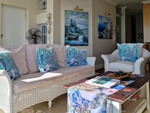 A living room with a wicker sofa, a white armchair, and a coffee table with a magazine. There are two paintings on the wall, a fan, and a door in the background. - The View (Cape Town)