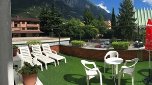 A terrace with white plastic chairs and tables, a red umbrella, and a view of a mountain and a building in the background. - Hotel San Marco BB (Darfo Boario Terme)