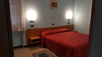 Una camera d'albergo con un letto singolo, un comodino, una piccola panca in legno, due luci a parete e un quadro incorniciato appeso al muro.