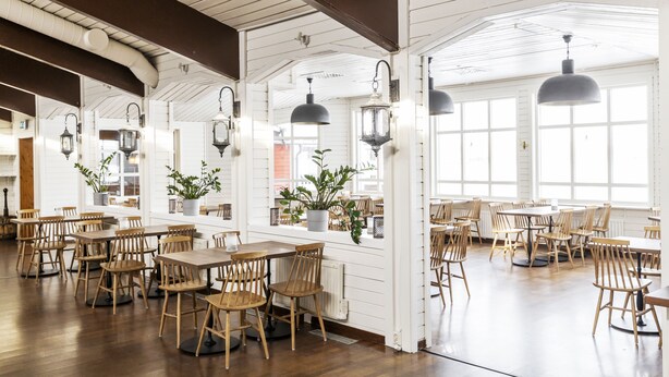 A spacious dining area with wooden tables and chairs, large windows, and hanging lantern-style lights. - Hotell Kraja (Arjeplog)