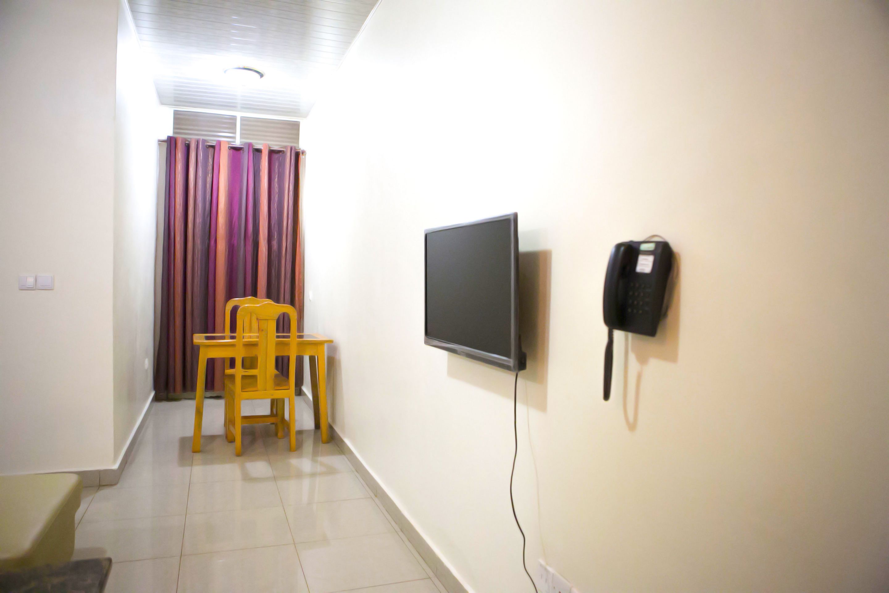 A room with a television mounted on the wall, a telephone, a chair, and a window with curtains.