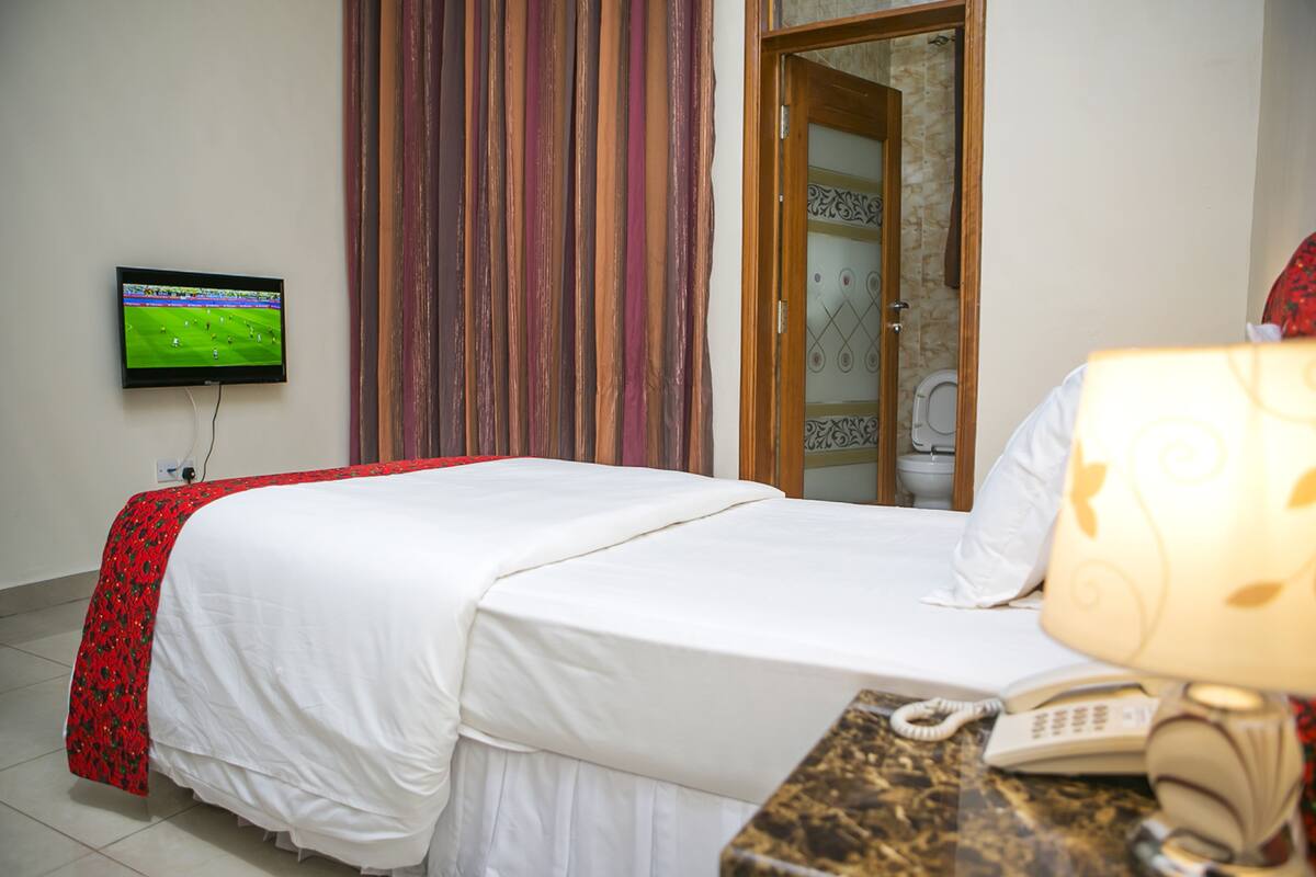 A hotel room with a bed, a TV mounted on the wall, a telephone, and a bathroom visible through an open door.