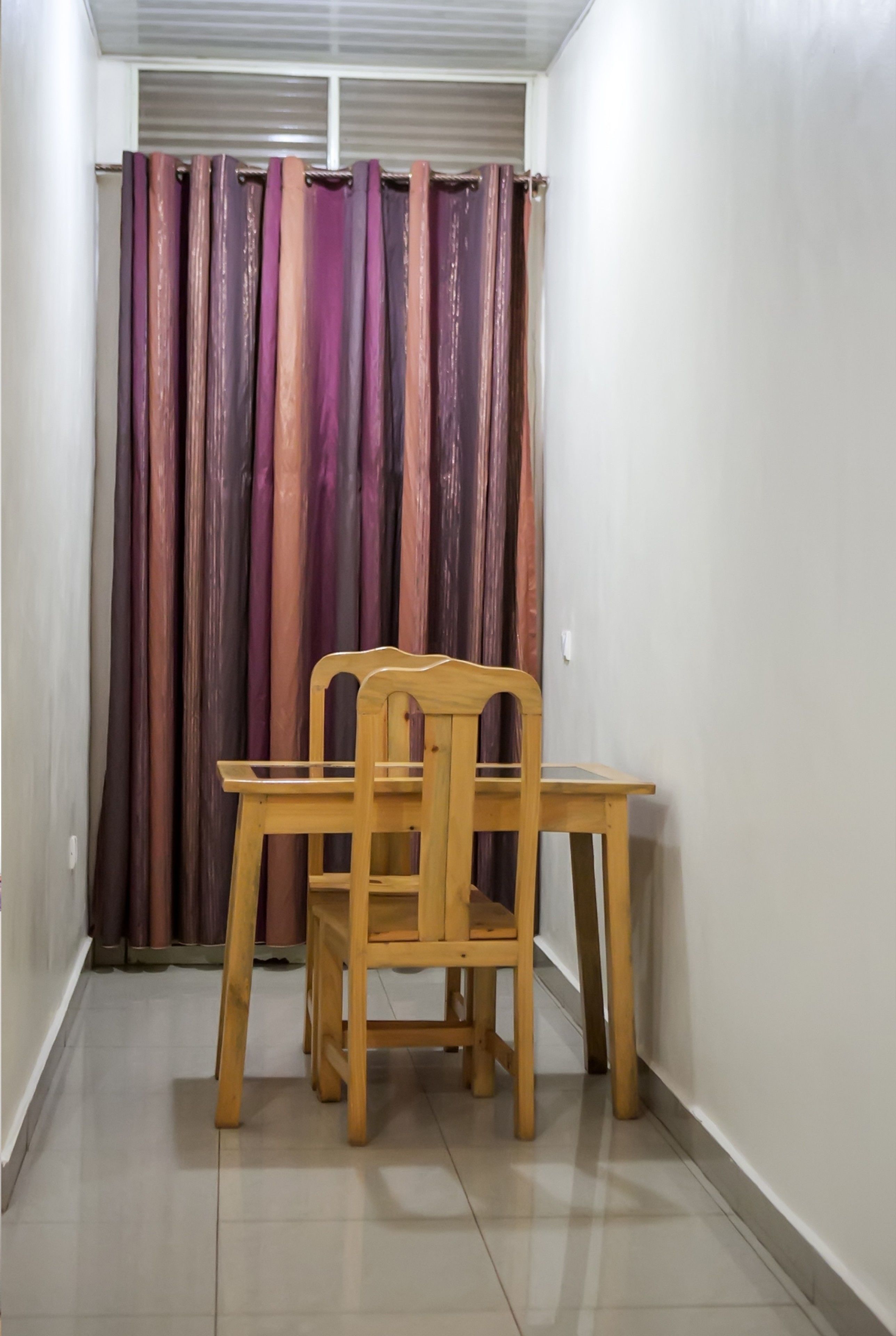 Un pasillo estrecho con una silla y una mesa de madera, una cortina y un piso de baldosas.