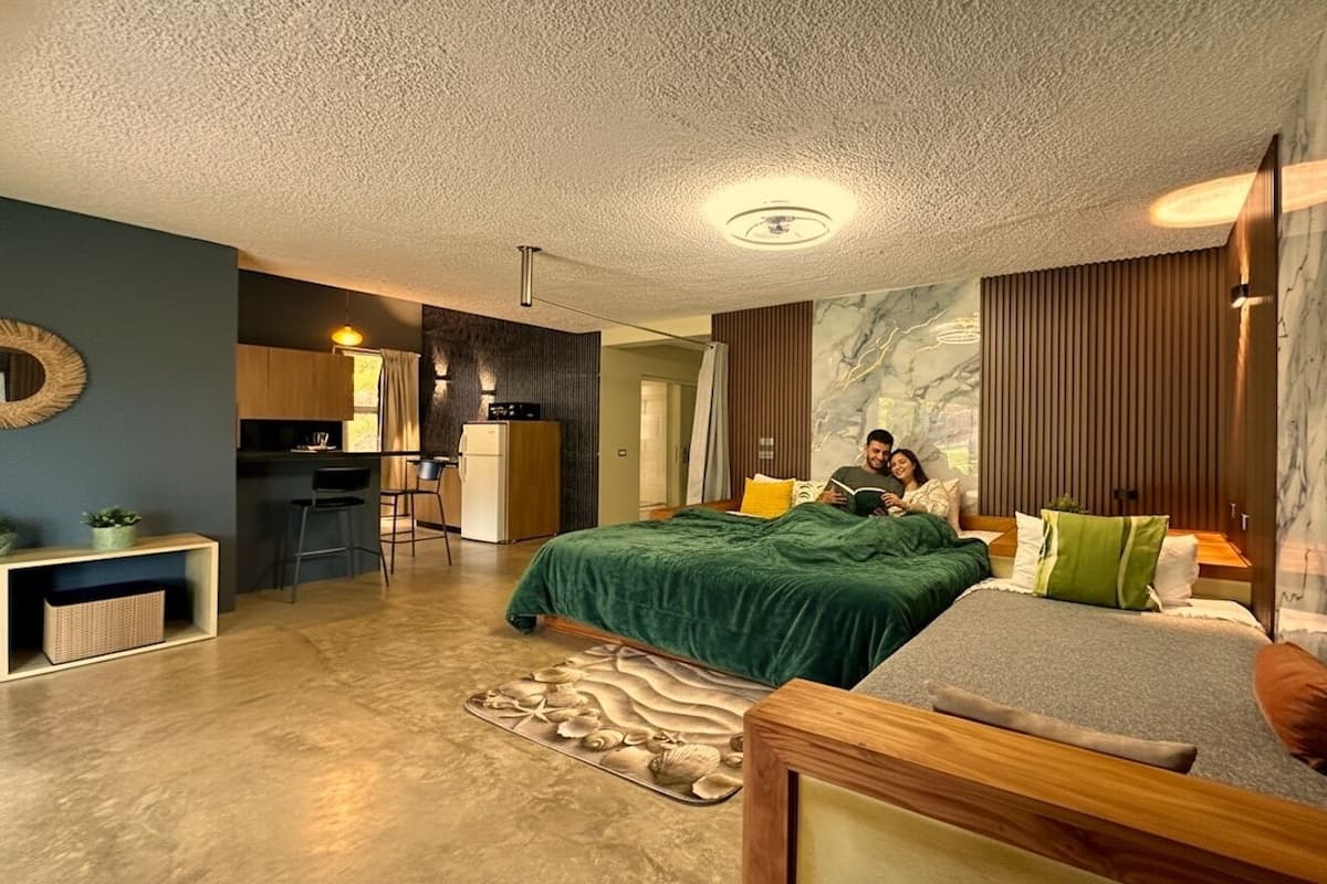 A modern bedroom with a large bed, a sofa, a desk, and a kitchen area.
