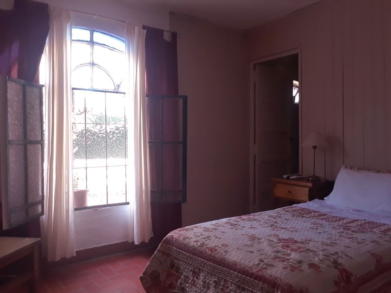 Un dormitorio con cama, mesita de noche, ventana con cortinas y puerta.
