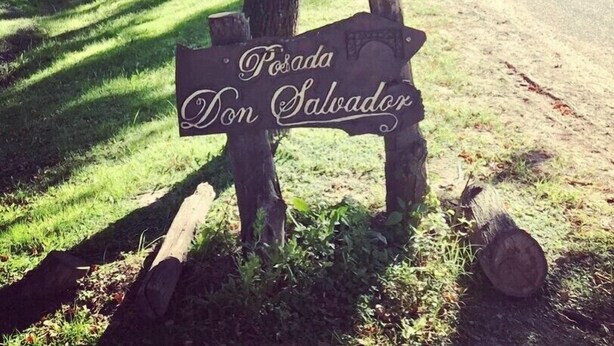 En rustik träskylt med texten ”Posada Don Salvador”, placerad i en naturskön miljö med träd och en grusväg.