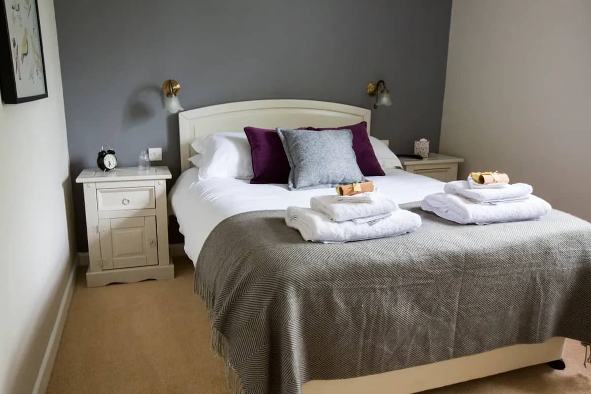 A neatly made bed with a grey blanket, two pillows, and a bedside table with a clock and a bottle.