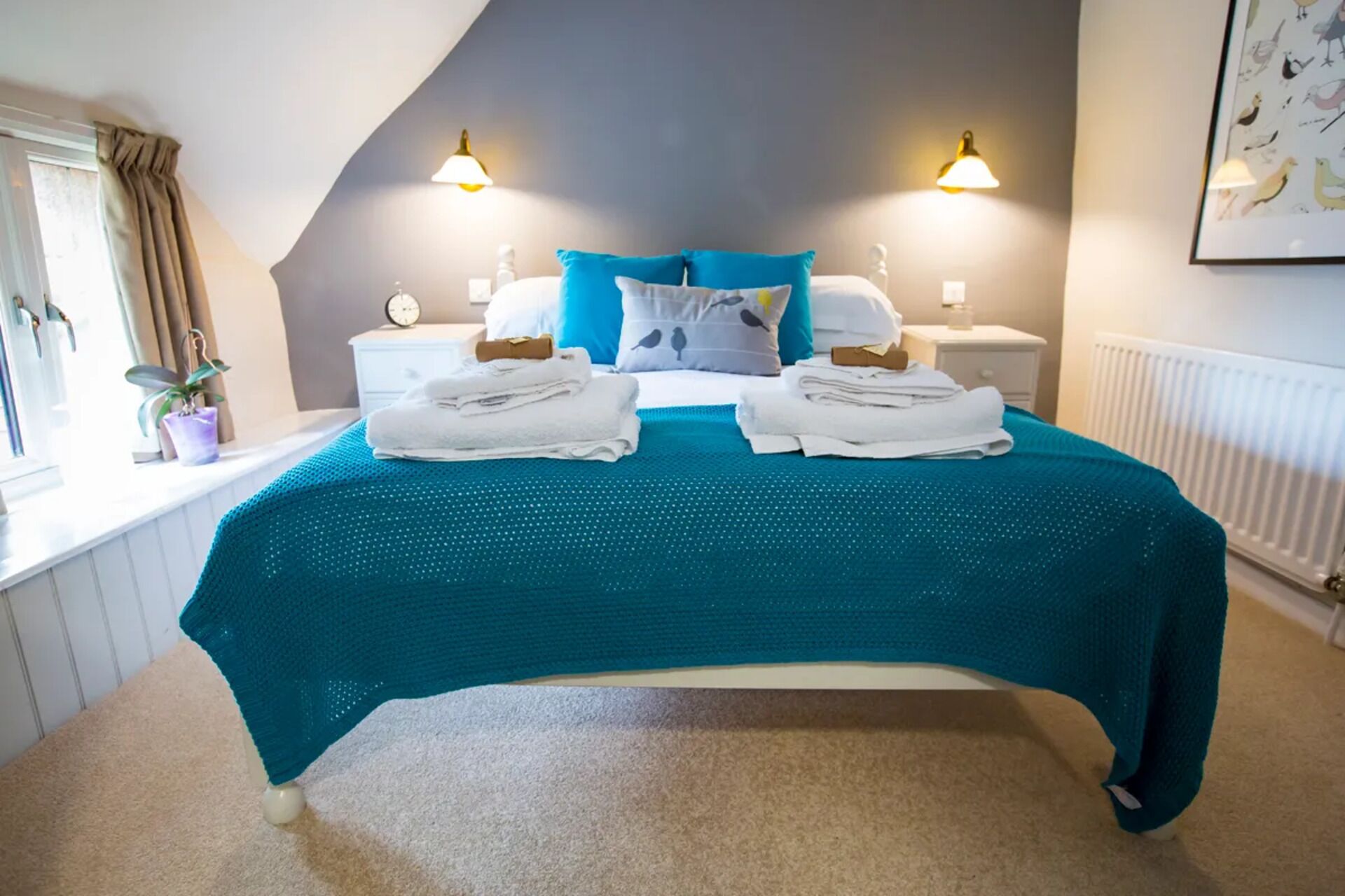 A bedroom with a bed, two bedside tables, a nightstand, a window with curtains, a radiator, and a wall-mounted light fixture.