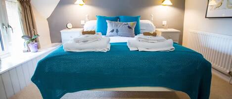 A bedroom with a bed, two bedside tables, a nightstand, a window with curtains, a radiator, and a wall-mounted light fixture.