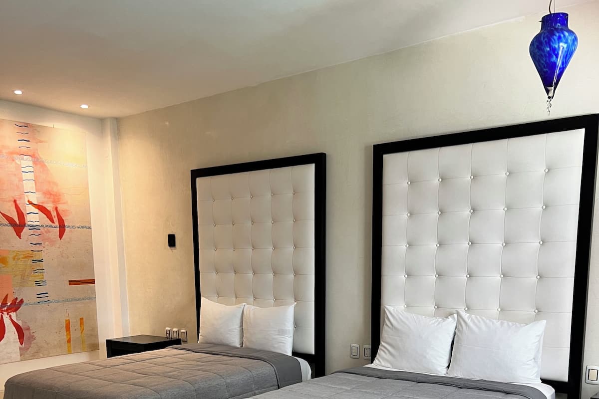 Une chambre d’hôtel moderne avec deux lits, une grande œuvre d’art au mur et un luminaire suspendu bleu.