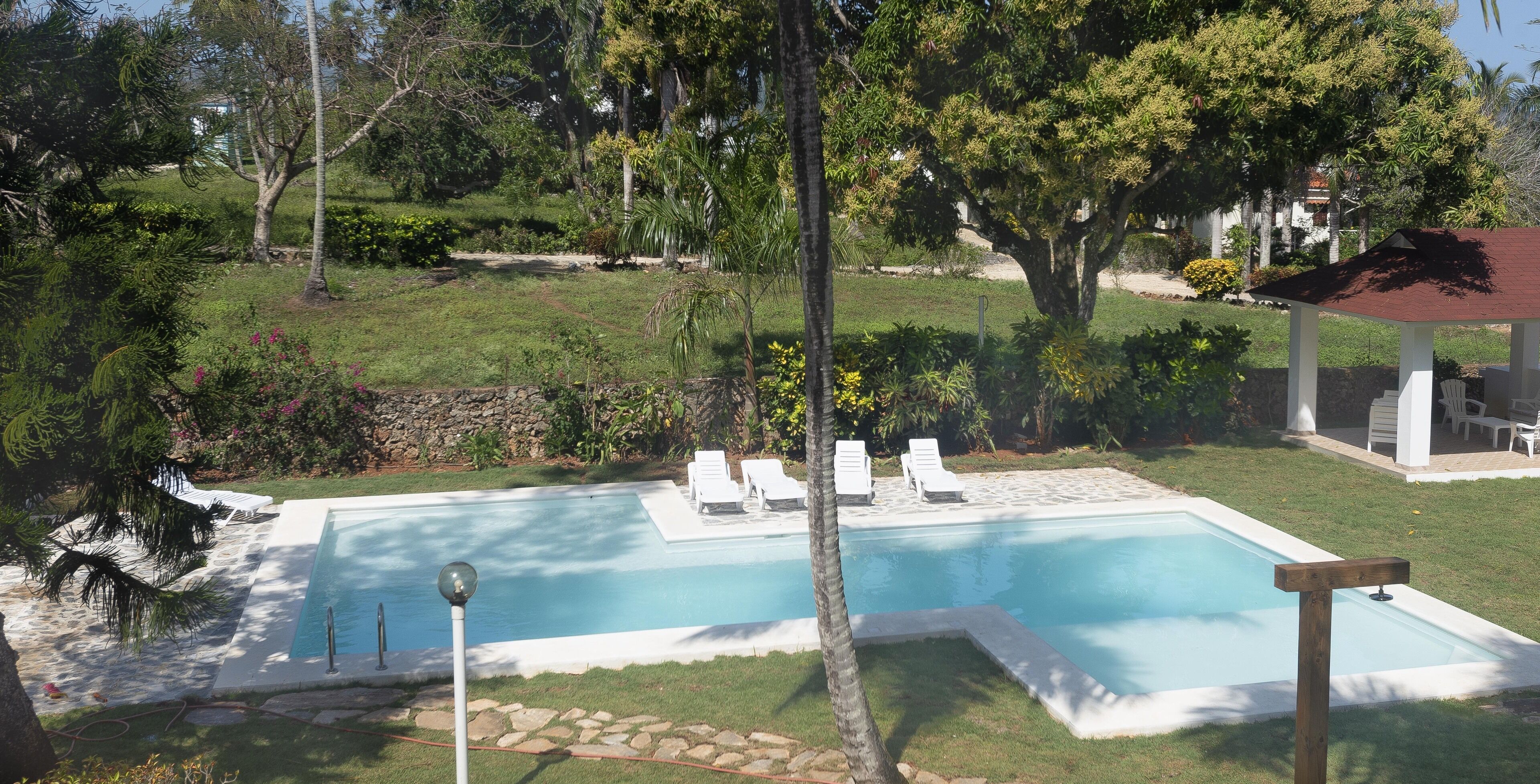 Una piscina rectangular con borde de piedra, rodeada por un césped cuidado y varios árboles. Se observan sillones de descanso y un pabellón al fondo.