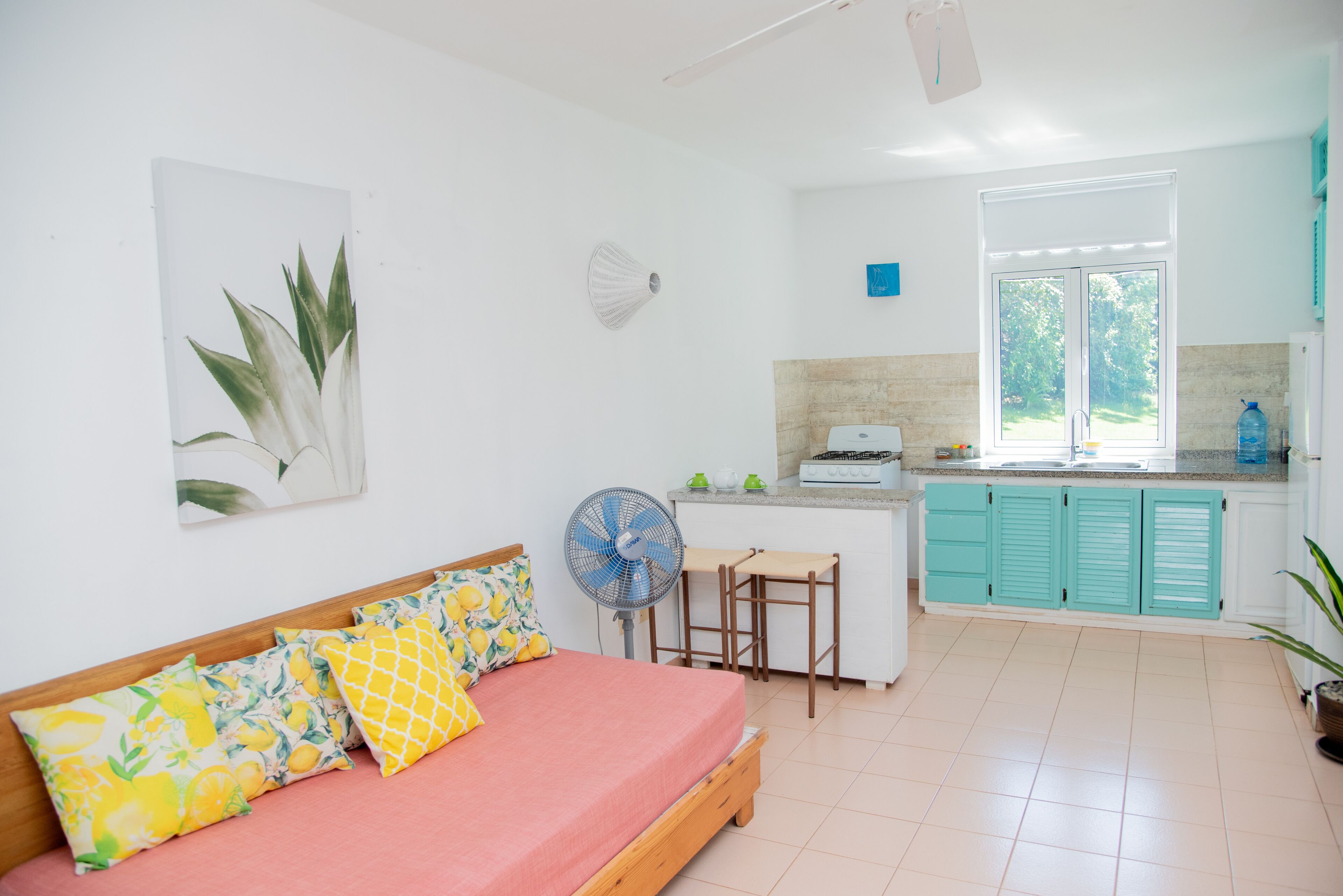 A bright room with a bed, a fan, a kitchen area, and a window.