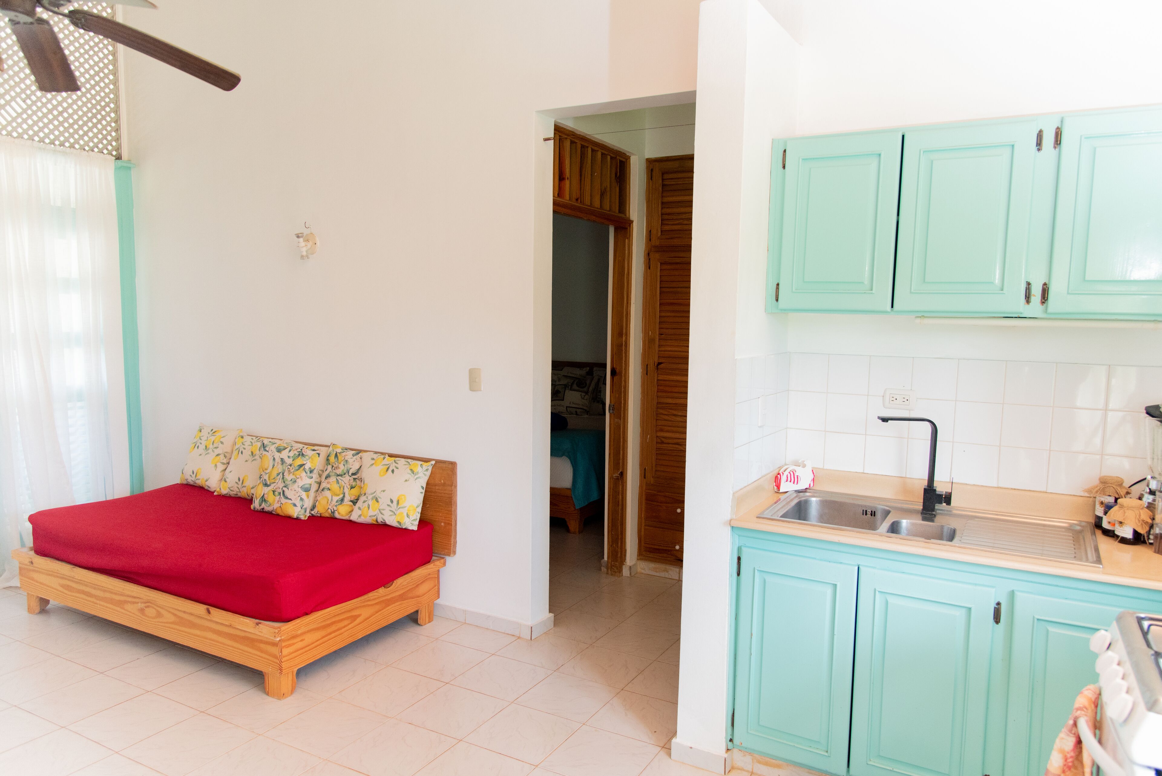 A bedroom with a wooden bed and a red blanket, a kitchenette with turquoise cabinets, and a bathroom visible through an open door.