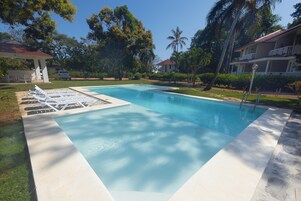 Piscina retangular com água azul límpida, rodeada por um deck branco. Há espreguiçadeiras e uma área de estar coberta com telhado vermelho. A piscina está situada numa área ajardinada com palmeiras e vegetação.
