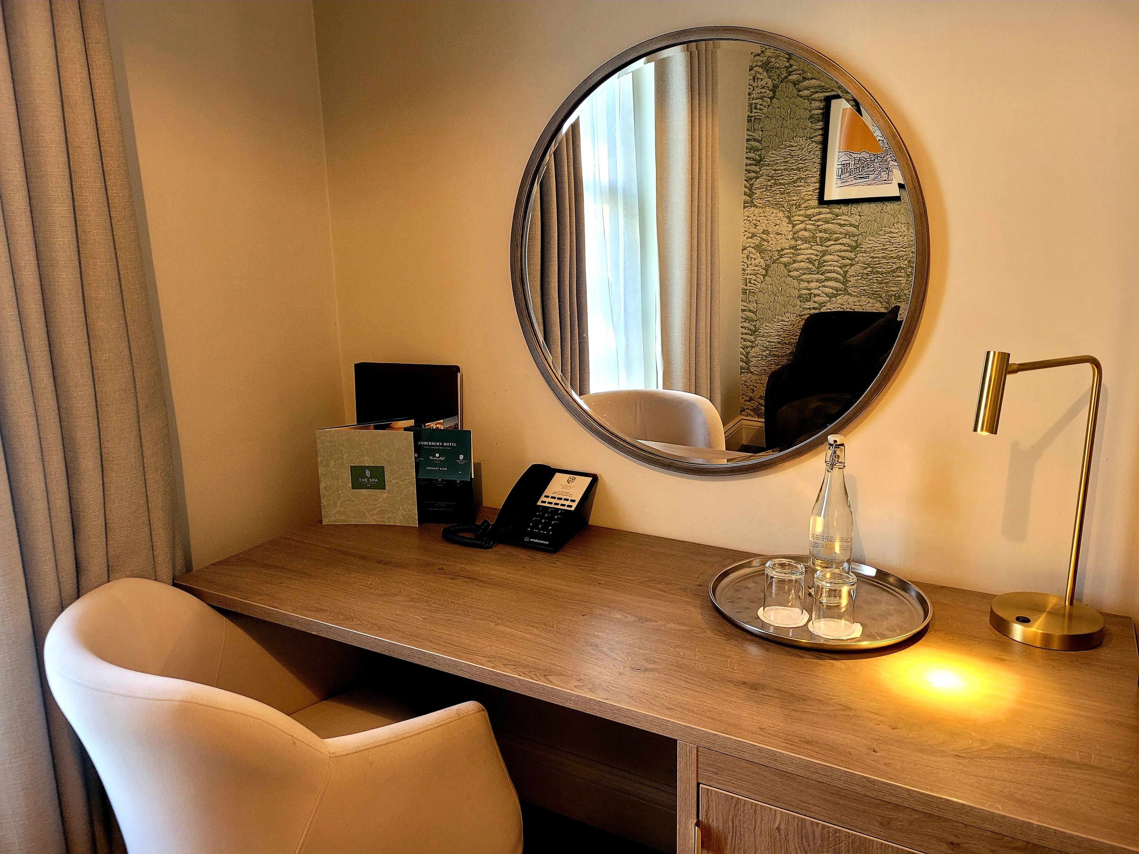 A hotel room with a wooden desk, a round mirror, a telephone, a bottle, and a tray with glasses.