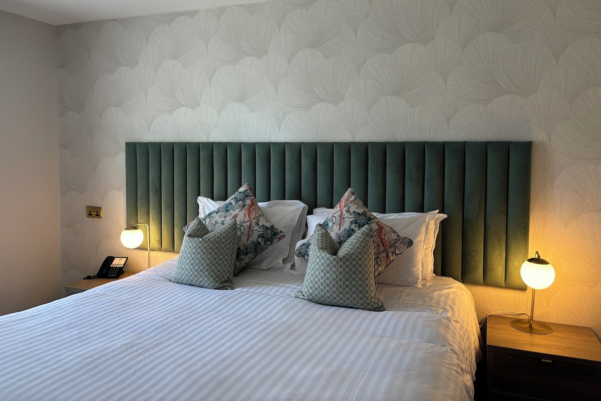 A neatly made bed with striped bedding, a green upholstered headboard, and decorative pillows. There are bedside lamps on either side of the bed, and a phone on the nightstand.