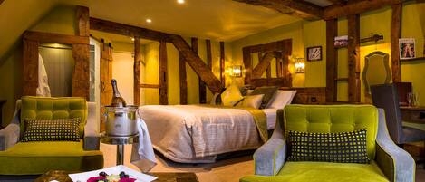 A cozy room with a bed, two green armchairs, a wooden ceiling, and a dining table with a fruit plate and cutlery.