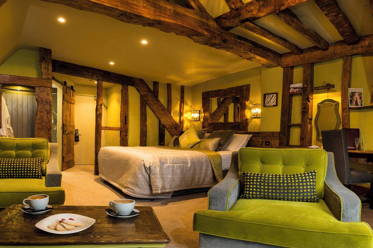 A cozy room with a bed, two green armchairs, a wooden coffee table with a plate of cookies, and a small table with a cup of coffee.