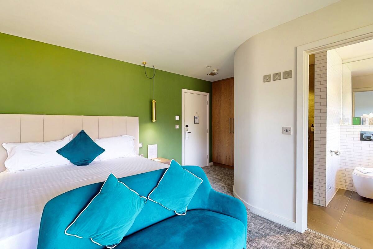 A hotel room with a bed, a teal sofa, a green wall, and a bathroom visible through an open door.