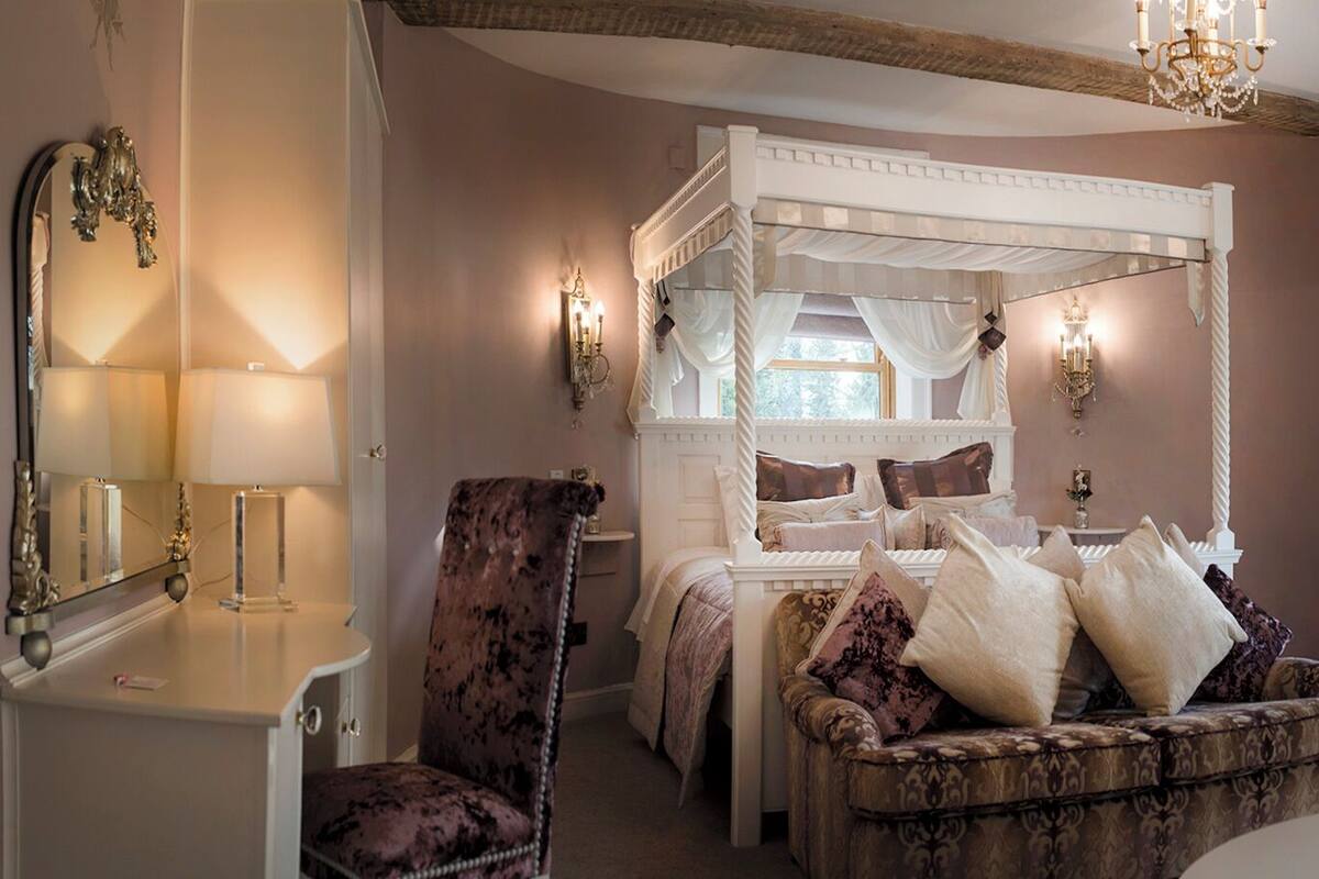 A bedroom with a four-poster bed, two upholstered chairs, a vanity with a mirror, and a vase with flowers.