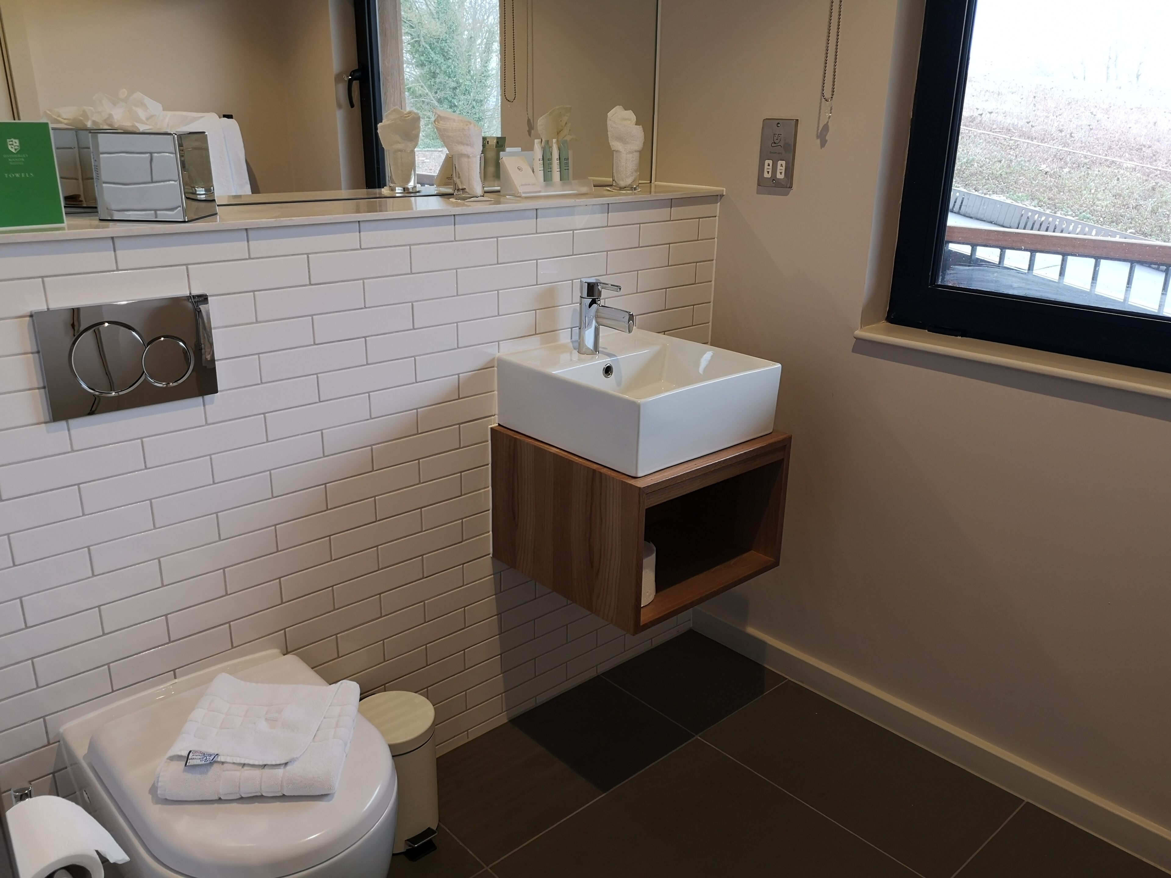 Une salle de bain moderne avec un mur recouvert de carreaux blancs, un meuble-lavabo en bois, des toilettes et une fenêtre donnant sur des arbres.