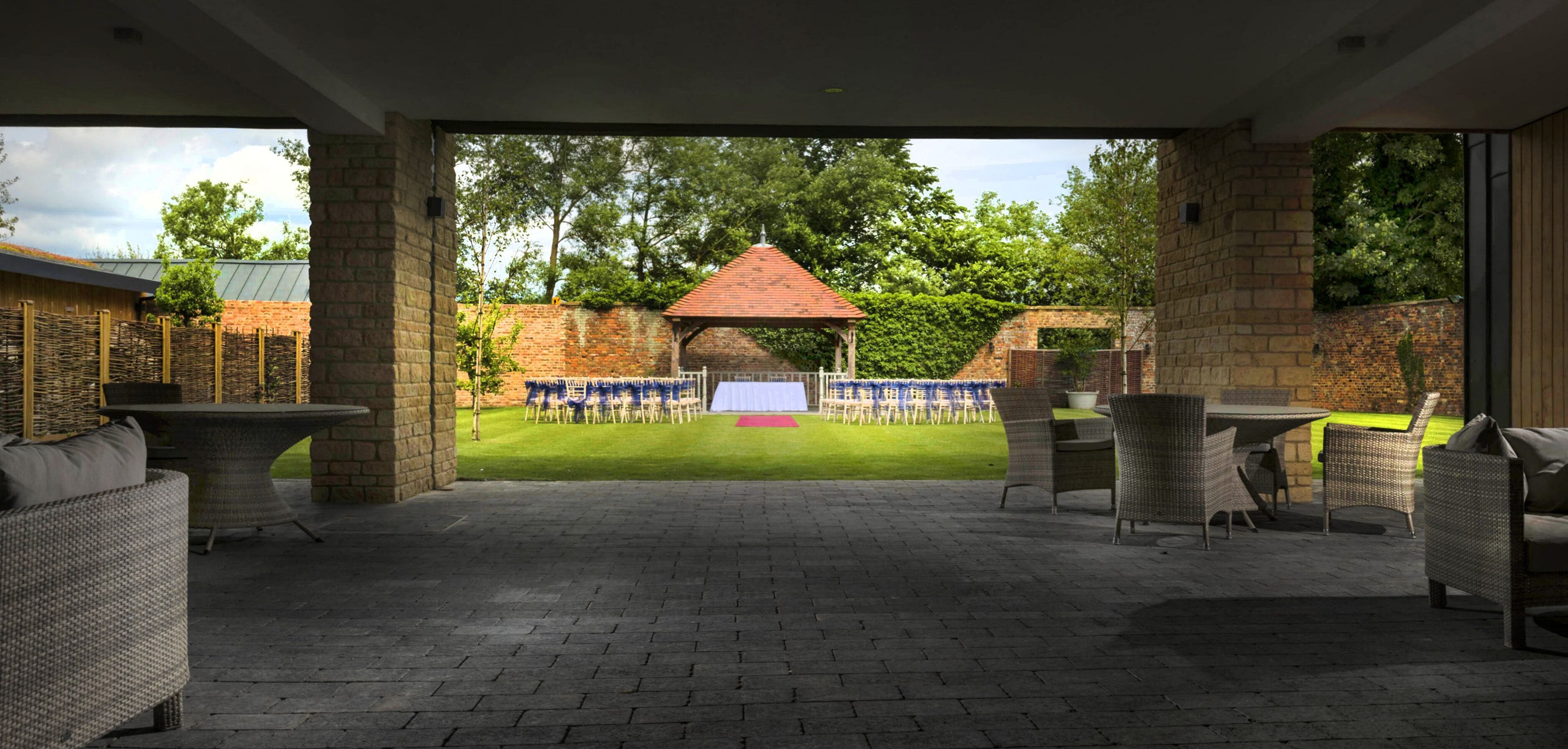 Ein Patio im Freien mit Korbmöbeln, einer Ziegelwand und Blick auf einen Garten sowie ein Gebäude mit rotem Dach.