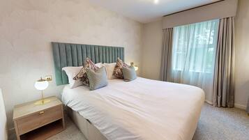 Cama bem arrumada com cabeceira estofada, mesa de cabeceira com abajur e uma janela grande com cortinas.