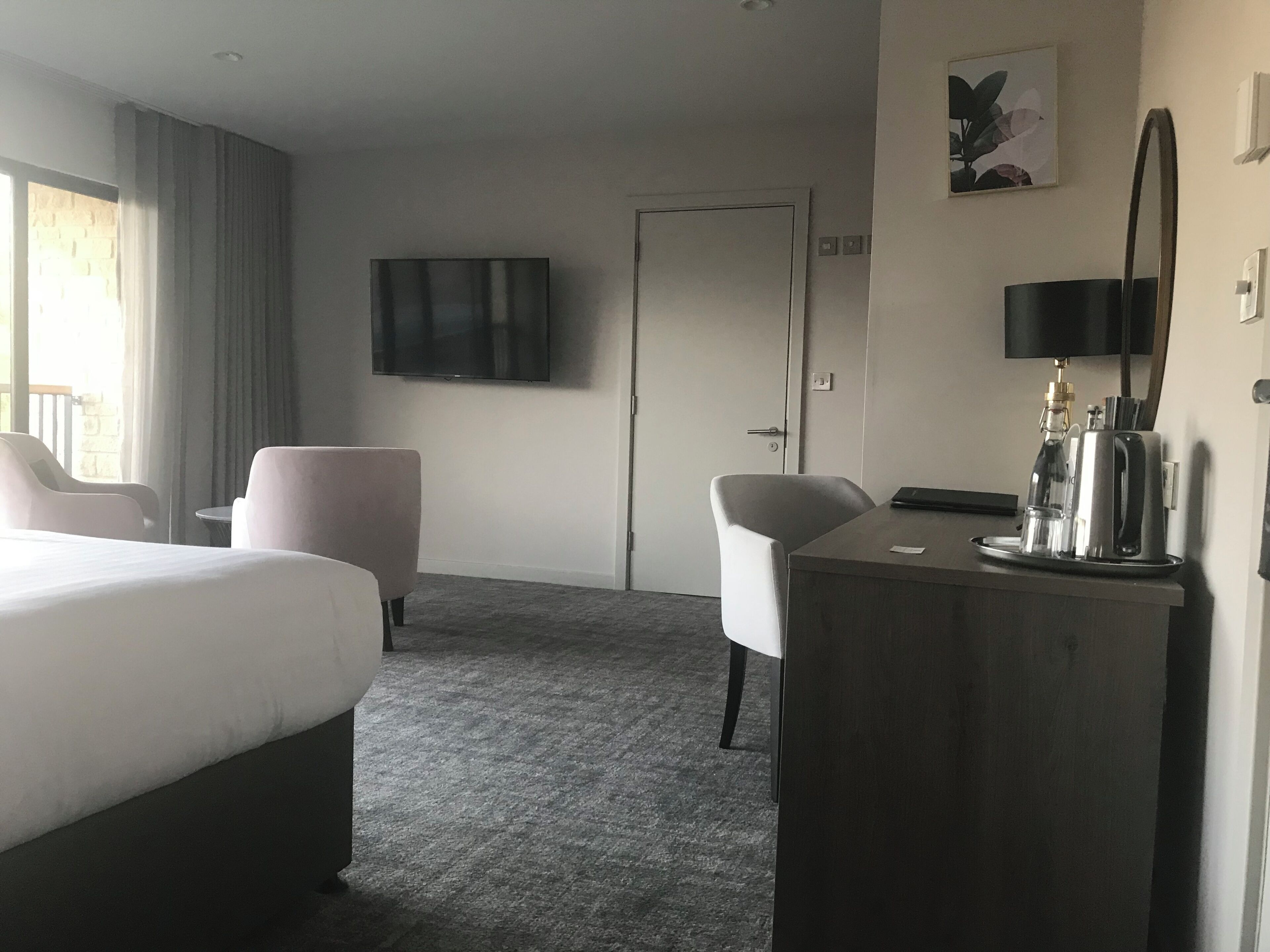 Hotellihuone, jossa on sänky, kaksi tuolia, työpöytä, kahvinkeitin, televisio ja ovi.